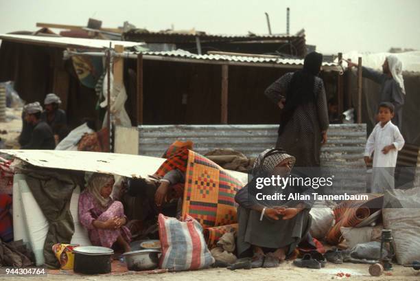 View of displaced men, women, and children as, surrounded by their belongings, they sit under and beside a makeshift shelter at a refugee camp during...