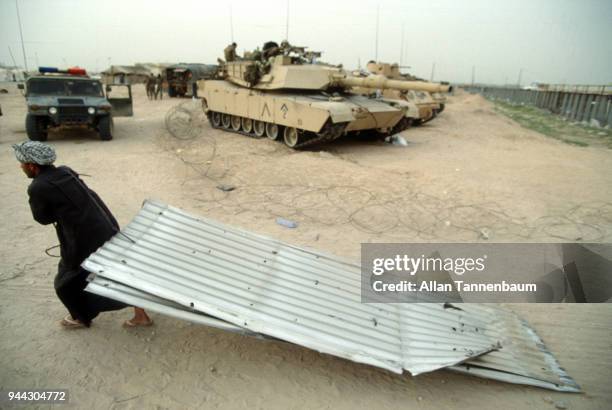 During the Gulf War, an Iraqi man drags a sheet of corrugated steel past a line of tanks at a refugee camp, Iraq, 1991.