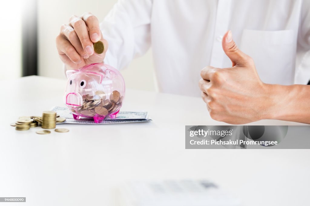 Midsection Of Businessman Putting Coin In Transparent Piggybank At Desk