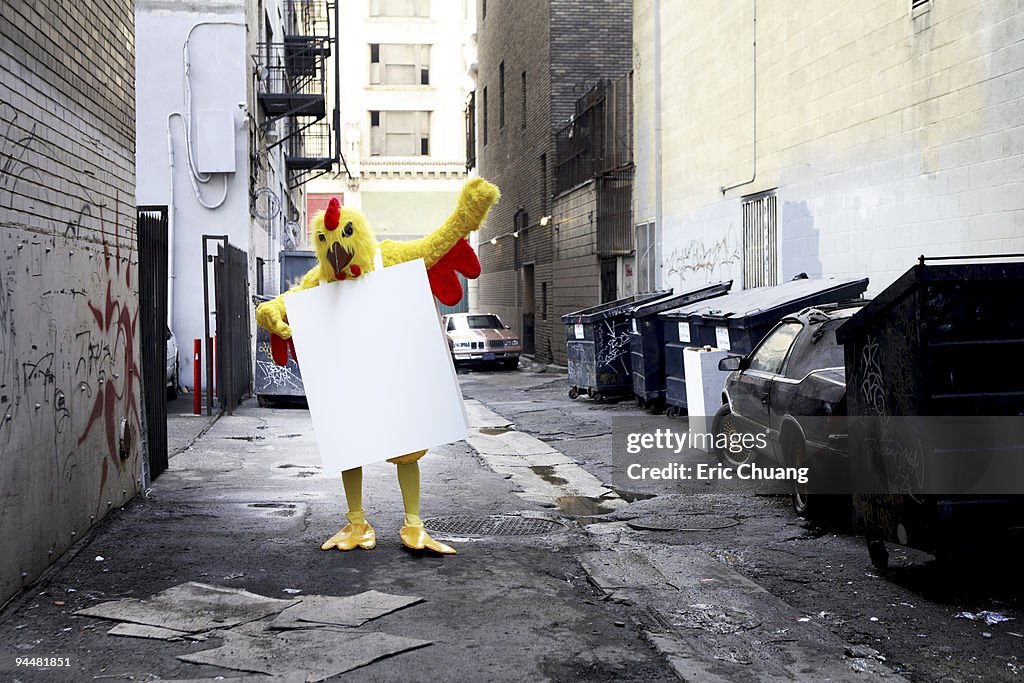 Person in chicken costume wearing signboard in alley