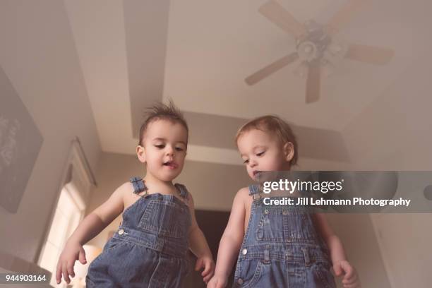 fraternal twin toddlers stand on a bed wearing bib overalls ready to jump - bib overalls stockfoto's en -beelden