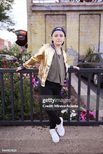 A young girl with cancer, wearing a gold jacket, posing in front of a metal gate.