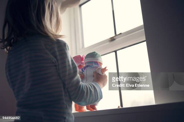 little girl in a dark room, holding two dolls and looking out of a window - looking through a doll house stock-fotos und bilder