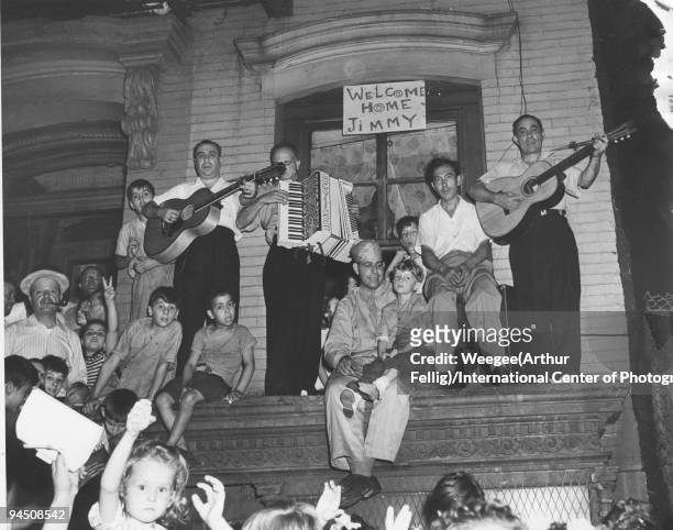 People standing in the sidewalk and on tenement steps; on a ledge sits a boy, Jimmy Di Maggio, on the lap of a man wearing a military uniform and a...