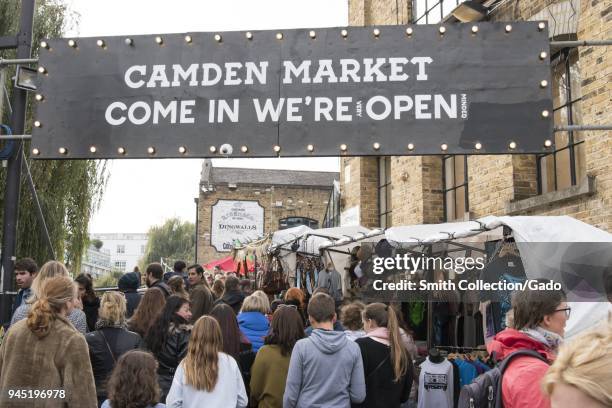 Crowds of shoppers, passing under an entrance sign reading "Camden Market Come in We're Open , " and walking on a pedestrian street, lined with...