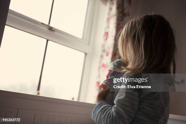 little girl in a dark room, holding a doll and looking out of a window - looking through a doll house photos et images de collection