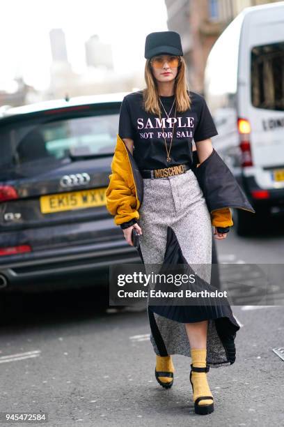 Guest wears a cap, a black t-shirt with a printed "Sample Not For Sale", a Moschino belt, an orange bomber jacket, a gray skirt, yellow socks, shoes,...