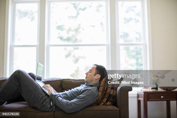 caucasian man working on a lap top while on a couch in his home. - 2000 teleworking stock pictures, royalty-free photos & images