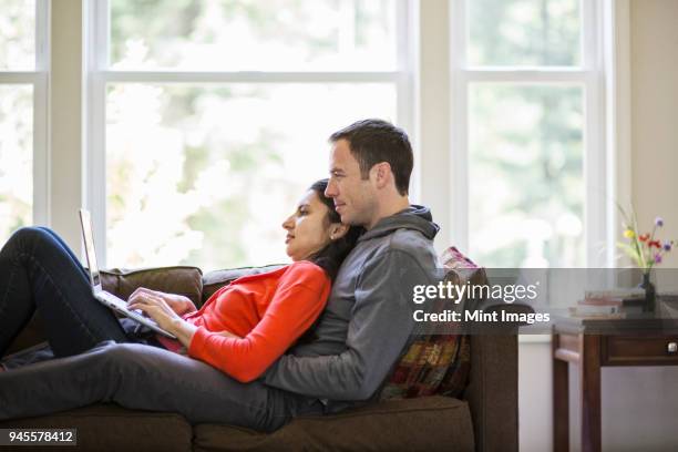 caucasian man and mixed race caucasian woman using a lap top computer while sitting on a couch in their home. - woman sitting top man stock pictures, royalty-free photos & images