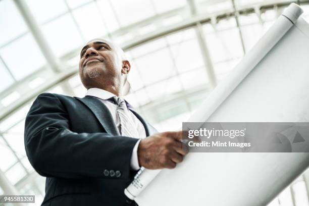 a black businessman holding plans for a new office space in a large glass covered walkway. - 異常角度 個照片及圖片檔