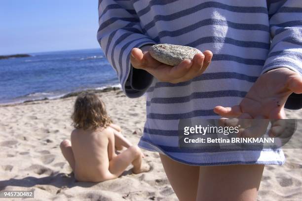 Un enfant tient un galet dans la main. Au fond, une fille de 4 ans est assise nue sur le sable d'une plage de l'Ile de Groix.