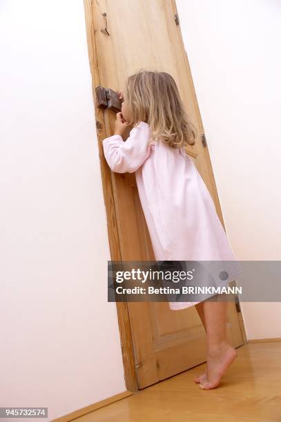 Une petite fille de 4 ans habillee en chemise de nuit regarde ? travers le trou d'une serrure d'une porte en bois.