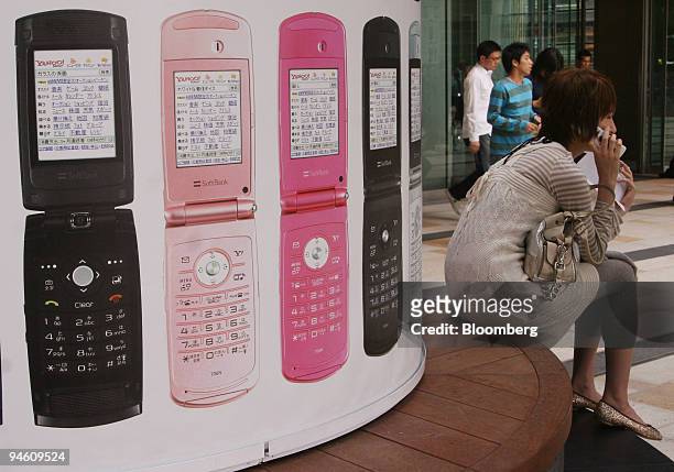 Woman uses her mobile phone as she sits by a pillar covered in Softbank Corp. Advertising in Tokyo, Japan, on Monday, May 7, 2007. Softbank Corp.,...