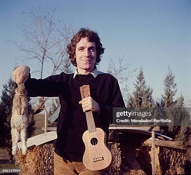 Italian singer-songwriter Rino Gaetano posing holding a rabbit by the ears and a ukulele in his hands. Photo shoot in the countryside. Italy, 1978