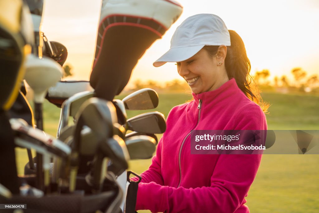 Mujer golfista asiendo clubes fuera de su bolsa de golf al atardecer.