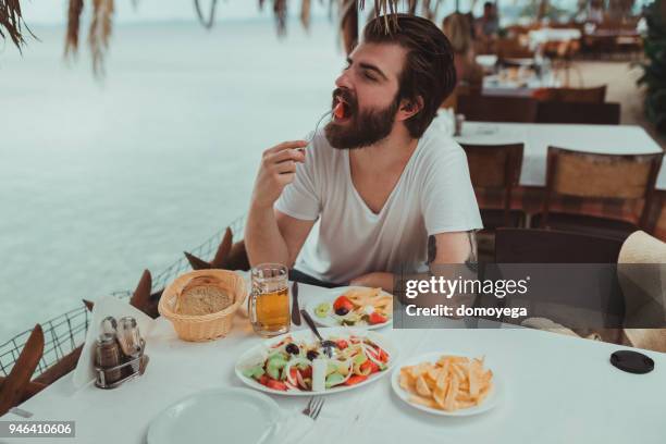 handsome bärtiger mann essen im restaurant nahe dem strand an einem sommertag - griechischer salat stock-fotos und bilder