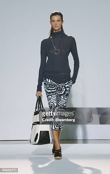 Model walks down the catwalk wearing a navy ribbed viscose roll neck over a navy rosette print cropped trouser and carrying black and white canvas...