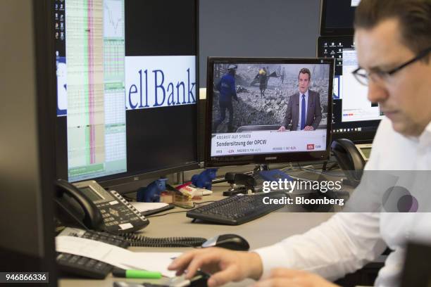 Television news report shows damage from military airstrikes on Syria as a trader monitors financial data inside the Frankfurt Stock Exchange,...