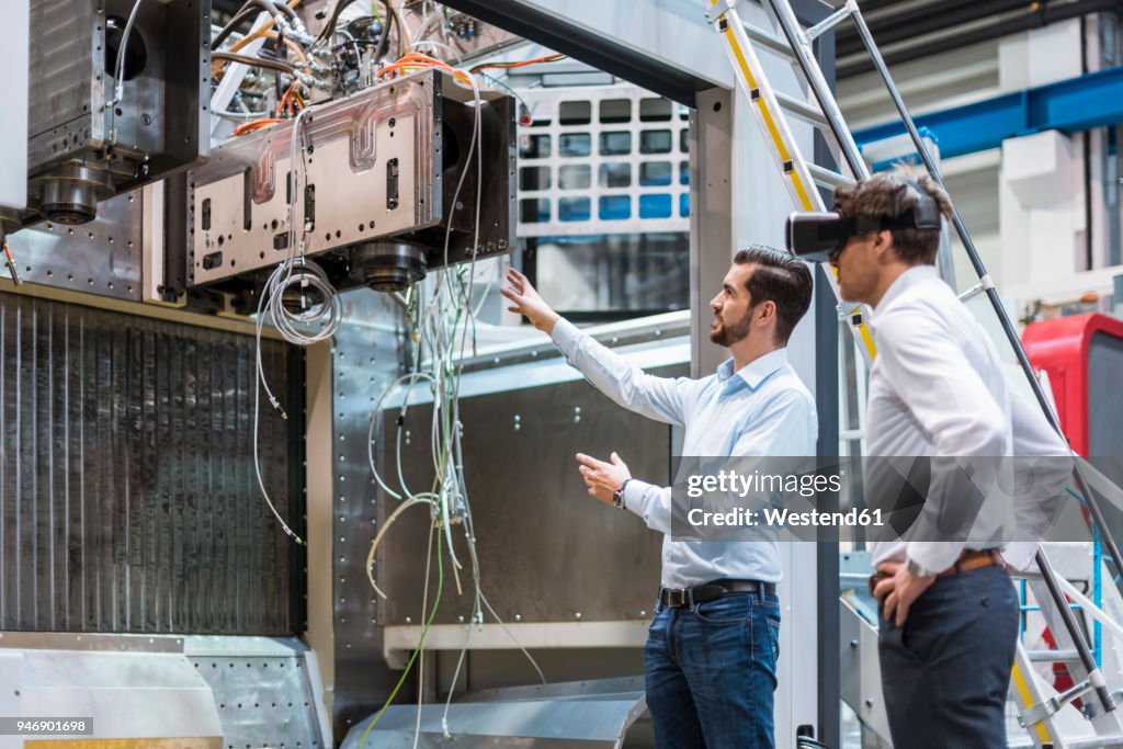 Man explaining machine to colleague wearing VR glasses in factory