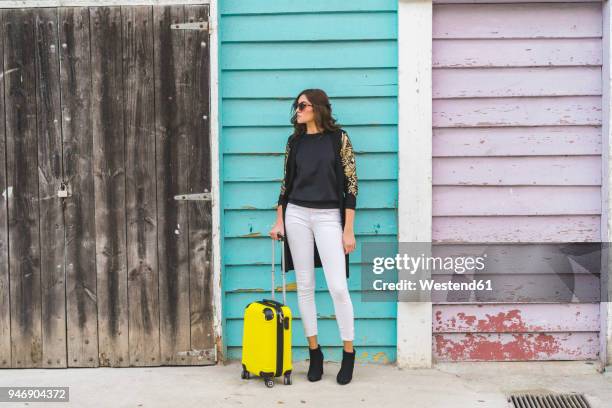 young woman with a yellow trolley bag waiting in front of wooden facade - luggage trolley stock-fotos und bilder