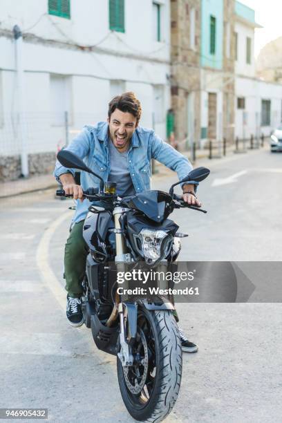 portrait of screaming young man sitting on motorbike pulling funny faces - moto humour photos et images de collection