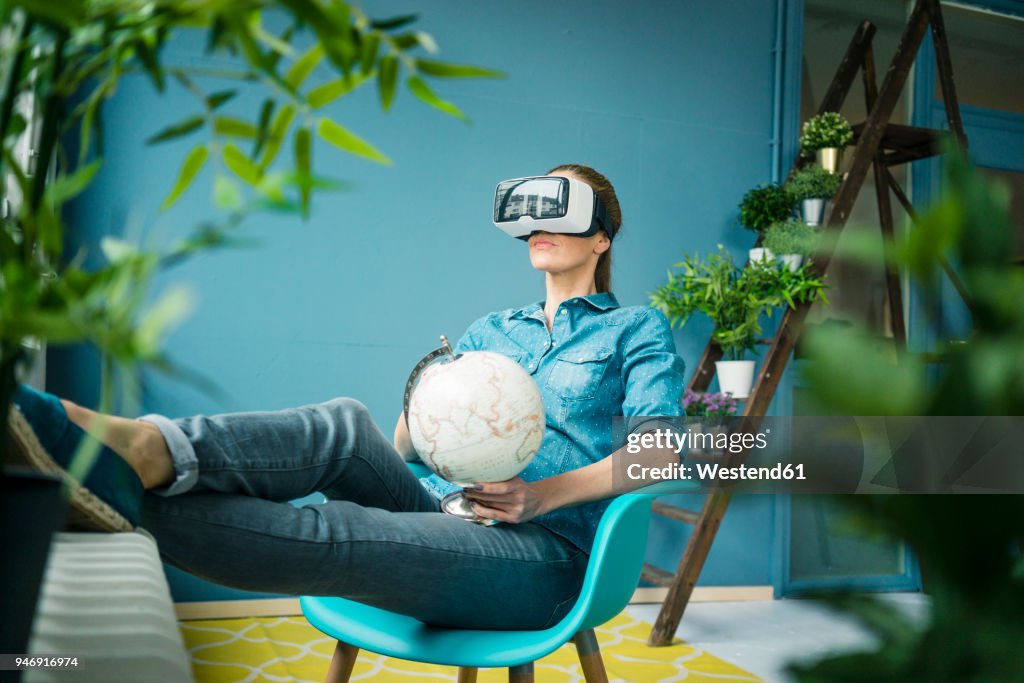 Beautiful woman sitting in her home, decorated with plants, holding globe, wearing VR glasses
