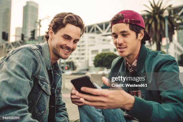 two smiling young men sharing cell phone outdoors - smartphone friends stock-fotos und bilder