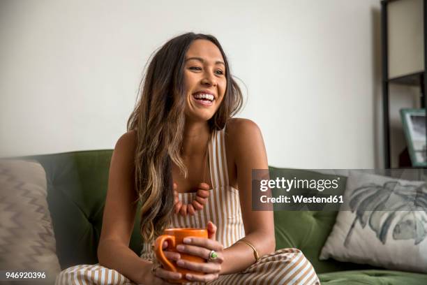 laughing young woman with coffee mug sitting on couch - lachende vrouw stockfoto's en -beelden