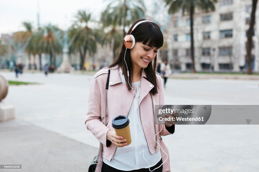 Spain, Barcelona, smiling woman with coffee, cell phone and headphones in the city