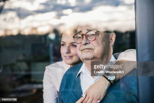 portrait of young woman embracing senior man at the window - care stock-fotos und bilder