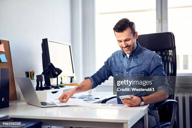 smiling man with smartphone and draft working at desk in office - employee mobile phone stock pictures, royalty-free photos & images