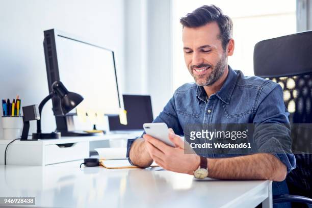 smiling man using cell phone at desk in office - business man looking at smart phone stock-fotos und bilder