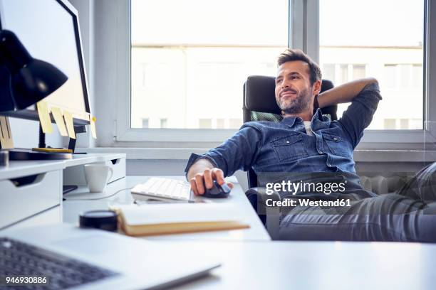 relaxed man sitting at desk in office looking at computer screen - bürostuhl stock-fotos und bilder