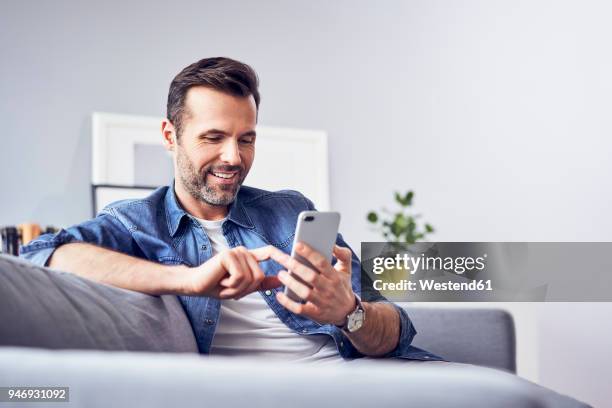 smiling man sitting on sofa using cell phone - movilidad fotografías e imágenes de stock