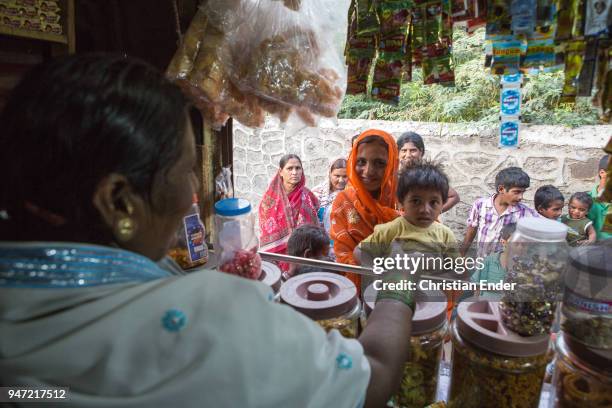 Several villagers standing in front of the counter of a small kiosk that is selling candy bars and other sweet treats in a poor neighbourhood on...