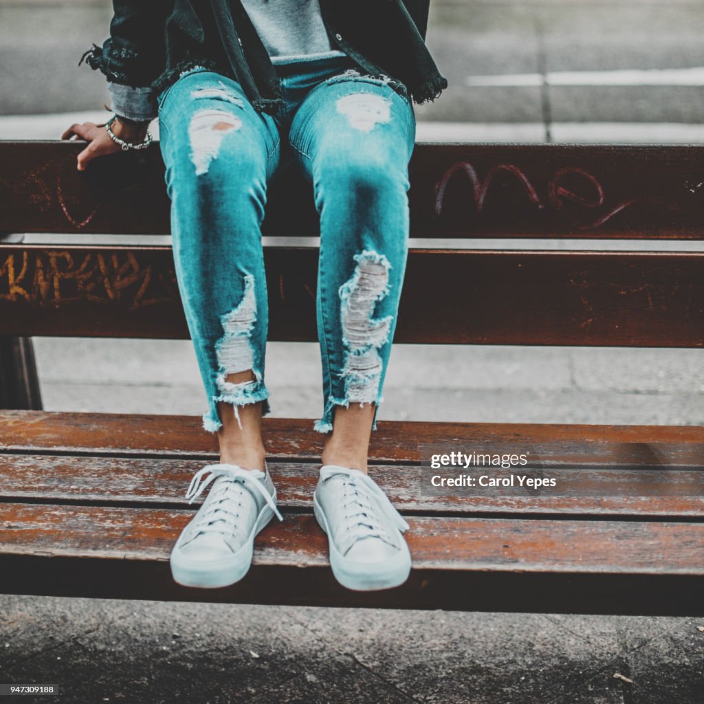 Teenage girl sitting on the park bench in denim  jeans sneakers