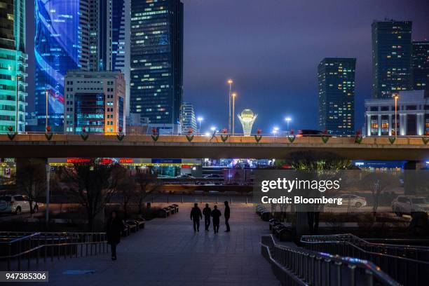 Pedestrians are silhouetted while walking a long a sidewalk as the Bayterak Tower is illuminated in the distance, center, at night in Astana,...