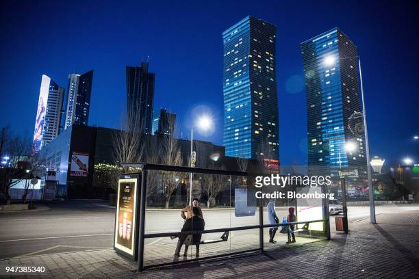 Passengers wait at a bus stop near the Northern Lights towers, right, standing among other residential and commercial buildings illuminated at night...