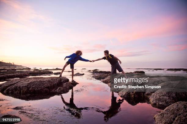 boys help each other across tidal pools at sunset - wasserrand stock-fotos und bilder