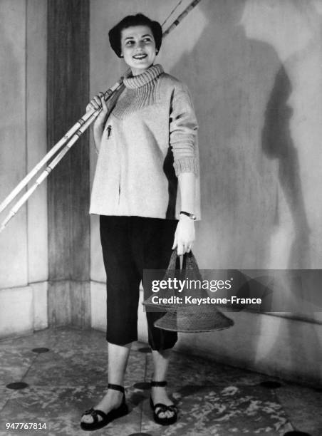 Une femme en tenue de pêche, son épuisette à la main et ses cannes à pêche sur l'épaule.