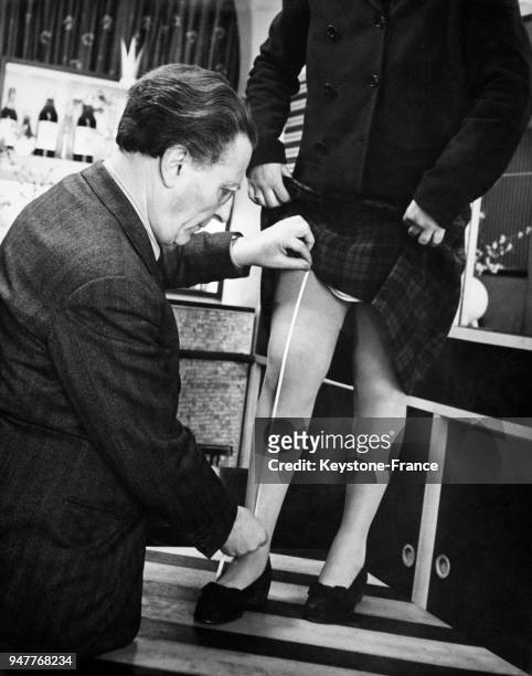 Un tailleur mesure la jambe d'une femme qui relève sa jupe, afin de lui confectionner une paire de collants sur mesure.