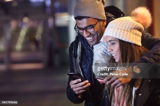 young couple at night, paying for something on phone with card - people diversity friends multi ethnic smiling indian ethnicity stock pictures, royalty-free photos & images