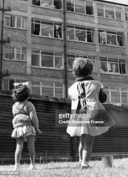 Deux petites filles dans un jardin d'enfants observent les fenêtres de l'usine voisine dans laquelle leurs mères travaillent, au Royaume-Uni.