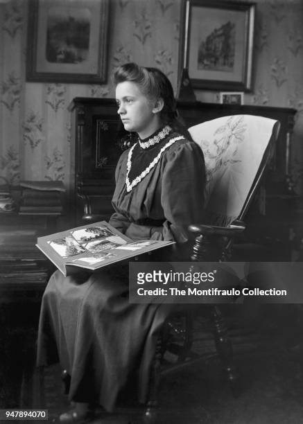 Young girl sitting on ornate wooden chair in living room holding a postcard album circa 1910.