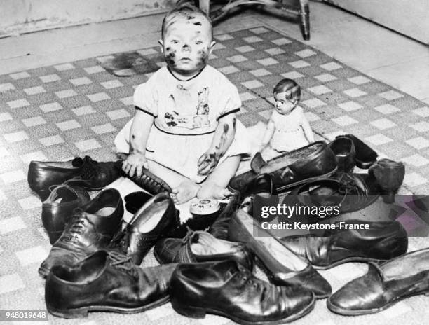 Un très jeune enfant, le visage maquillé de cirage, s'amuse à cirer des chaussures rassemblées devant lui, circa 1930.