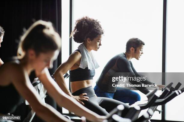 woman with towel around neck riding stationary bike during cycling class in fitness studio - heimtrainer stock-fotos und bilder
