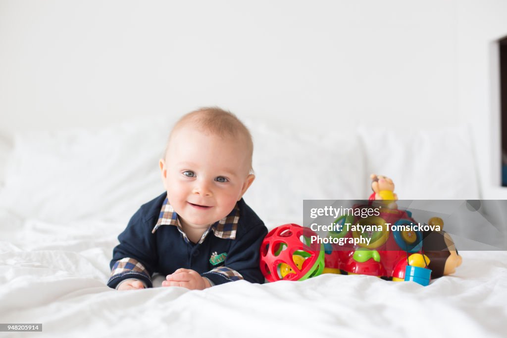 Sweet toddler boy, playing with colorful toys in bed, daytime. Childhood happiness concept, early development toys