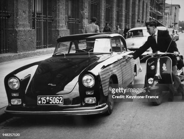Une homme en vespa regarde une voiture garée dont les côtés sont recouverts d'une résine la protégeant des rayures, à Munich, Allemagne en avril 1935.
