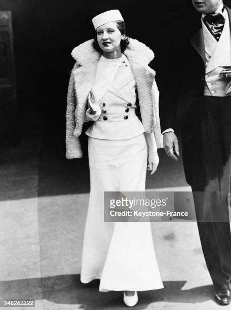Femme élégante portant un fez et vêtue d'une robe évasée dans le bas et un gilet seyant fermé par de gros boutons, à Ascot, Royaume-Uni en 1932.