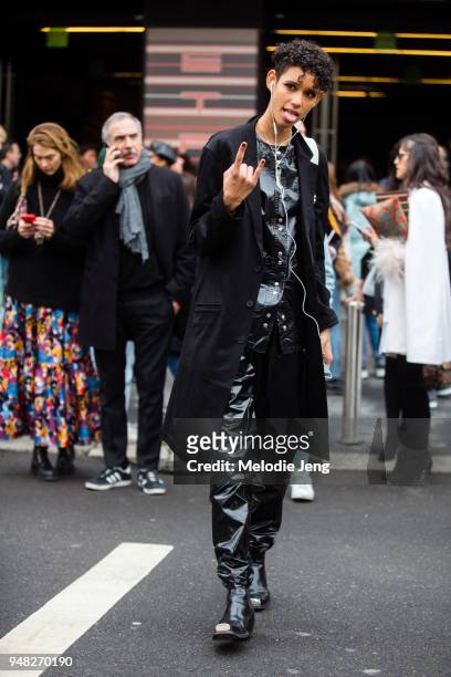 Model Dilone throws a hand signal and wears a black jacket over a latex black button up top and pants, and black leather Calvin Klein CK205W29NYC...